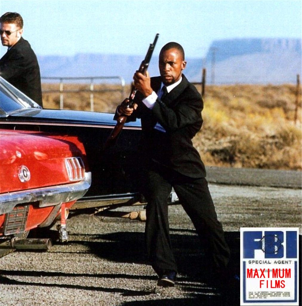Sensei San Ndlovu in a suit with a gun beside a red car, on the set of IBIS